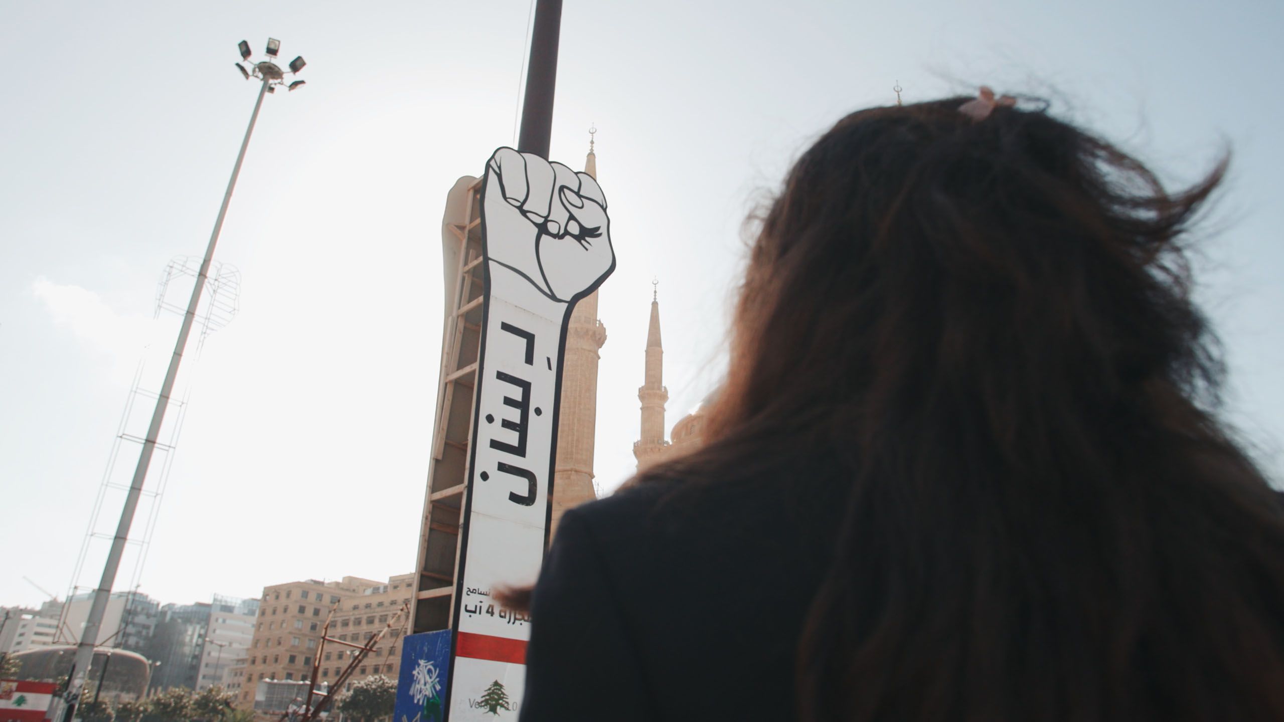 Eine Aktivistin steht vor einem großen Protestschild mit einer erhobenen Faust und arabischem Text in Beirut, ein zentrales Bild aus dem Libanon-Teil der Doku „Women Leading Protests“.