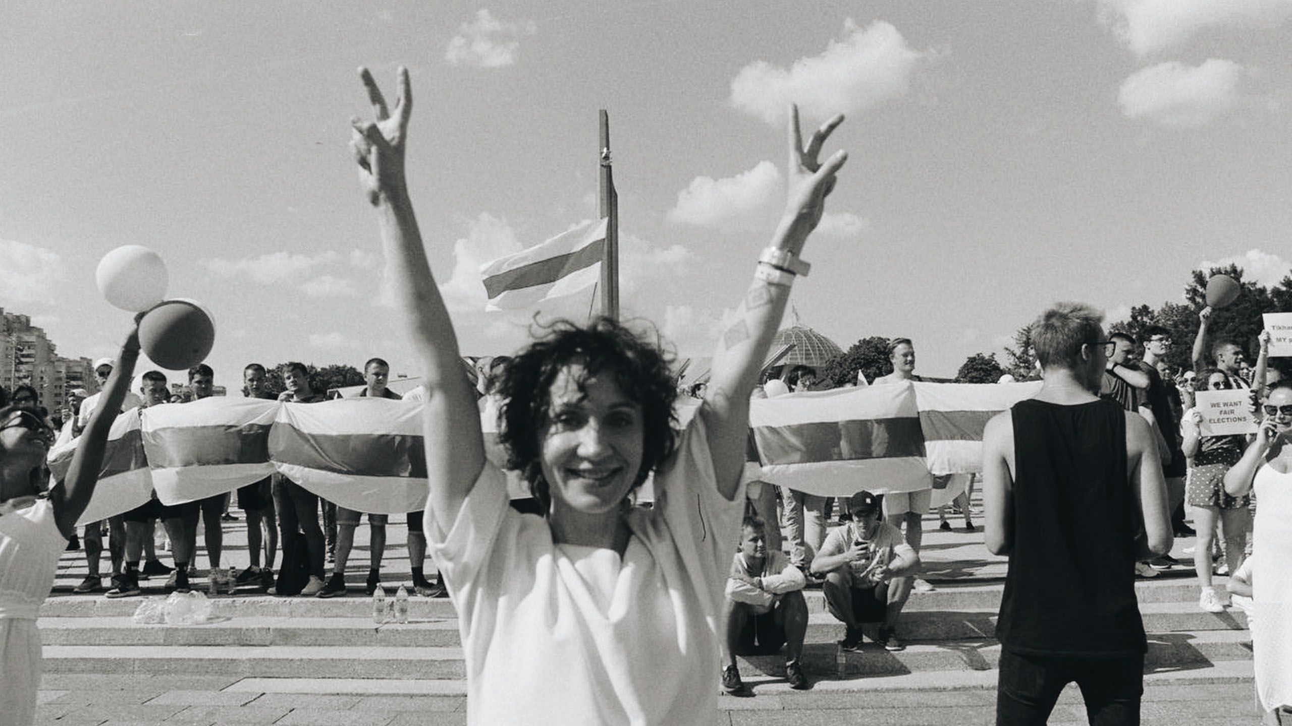 Ein kraftvolles Schwarz-Weiß-Standbild einer Aktivistin in Belarus, die mit einem hoffnungsvollen Lächeln während der Massenproteste für faire Wahlen das Victory-Zeichen zeigt, aus dem Film „Women Leading Protests“.
