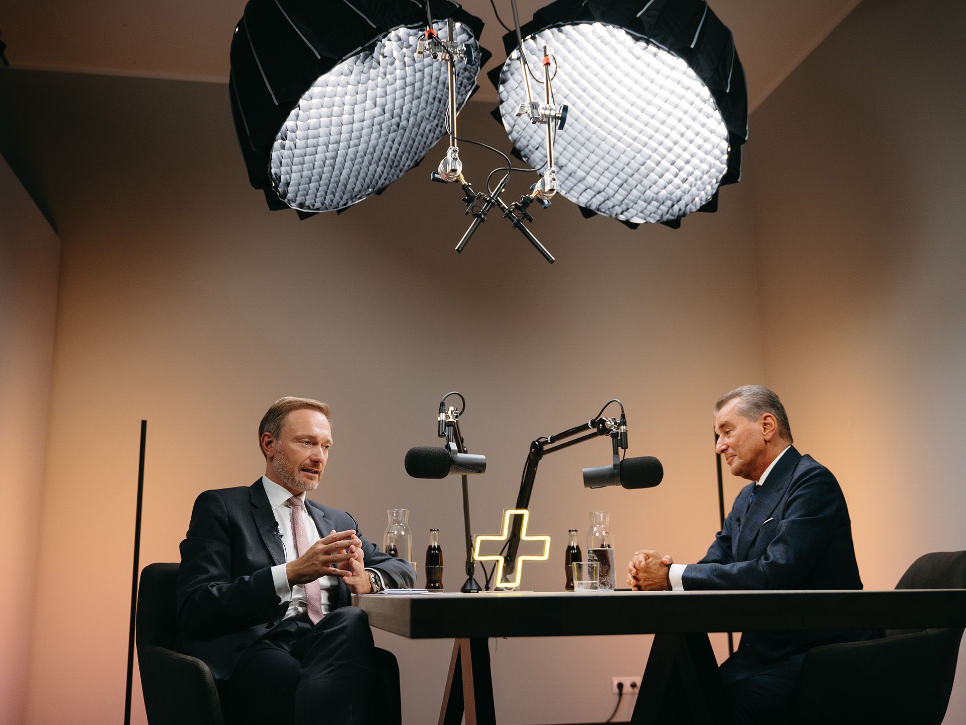 Eine Frontalansicht, die Christian Lindner im Interview mit einem Gast für den 'CL+' Podcast zeigt. Die Aufnahme hebt das professionelle Studio-Setup hervor, inklusive Mikrofonen und dem maßgefertigten Neon-Logo, alles produziert von Visionary Berlin